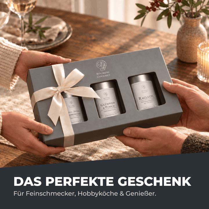 Zwei Personen überreichen sich die graue Boomers-Gourmet-Geschenkbox mit cremefarbener Schleife und drei Gewürzdosen darin